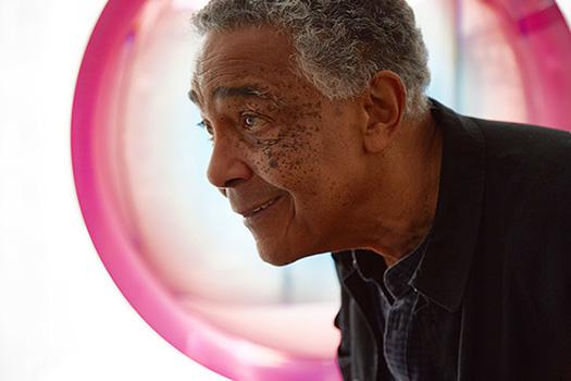 Man in foreground wearing black shirt looks upward, a circular translucent pink sculpture is in the background.