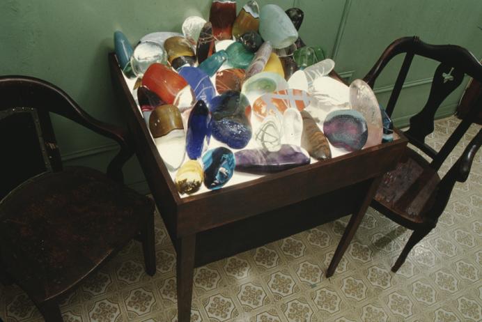 Arlan Huang, "100 Smooth Stones for Grandfather," 1994 Photo of an installation of large multicolored, smooth, transparent rock-shapes, made of glass, on a light table with chairs.