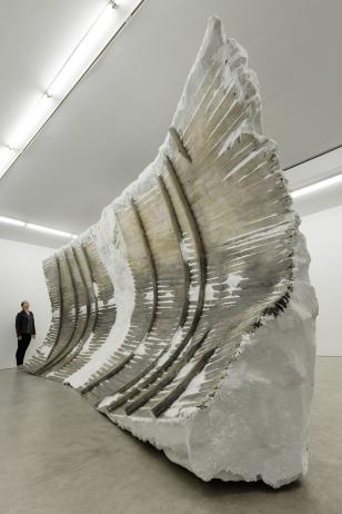 Photograph of a large curved, white sculpture with embedded wooden planks. A woman is standing at one end of the sculpture. 