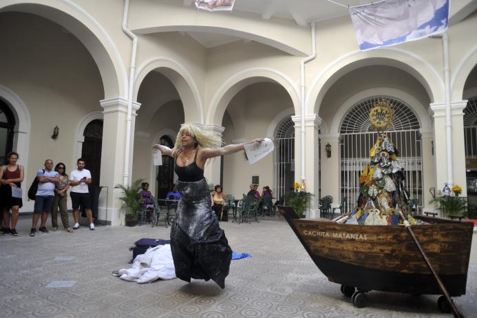 Photo of a woman dancing in a courtyard, dressed in black, with blond hair. A row boat with colorful flags is next to the figure