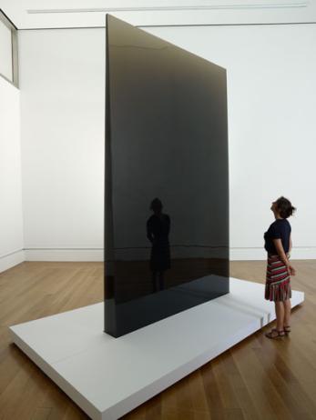 Photo of a large dark gray reflective and transparent triangular column, on a white plinth. A woman stands in front.  