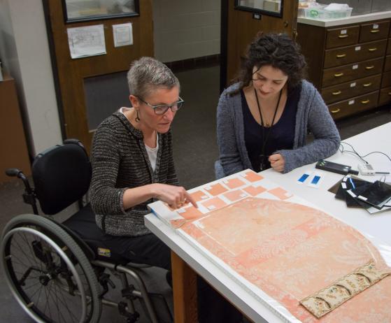 Photo of Wickens, seated in a wheelchair, teaching Murtore about light degradation and color-matching in textile conservation.