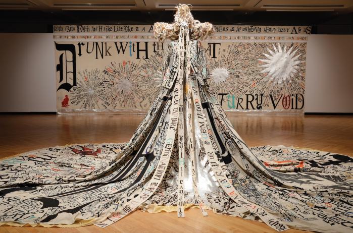 Installation photo of elaborate beige costume with black calligraphy on a mannequin in front of a hanging textile. 