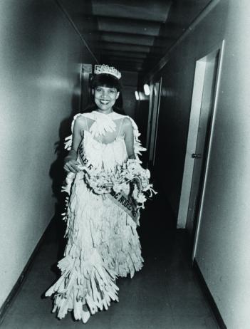 Photo of the artist dressed as a beauty queen, wearing a tiara, sash and gown made of gloves. She walks down a corridor smiling.