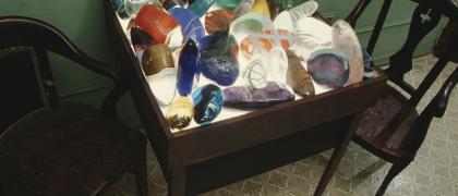 Photo of an installation of large multicolored, smooth, transparent rock-shapes, made of glass, on a light table with chairs.