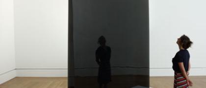 Photo of a large dark gray reflective and transparent triangular column, on a white plinth. A woman stands in front.  