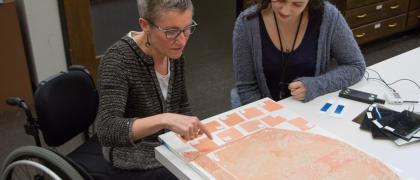 Photo of Wickens, seated in a wheelchair, teaching Murtore about light degradation and color-matching in textile conservation.