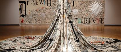 Installation photo of elaborate beige costume with black calligraphy on a mannequin in front of a hanging textile. 