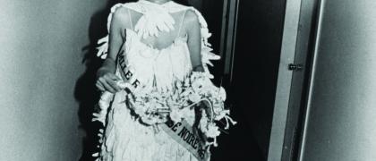 Photo of the artist dressed as a beauty queen, wearing a tiara, sash and gown made of gloves. She walks down a corridor smiling.