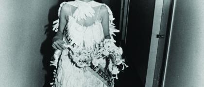 Lorraine O’Grady performing "Mlle. Bourgeoise Noire leaves the safety of home," 1980-83, printed 2009. Photo of the artist dressed as a beauty queen, wearing a tiara, sash and gown made of gloves. She walks down a corridor smiling.